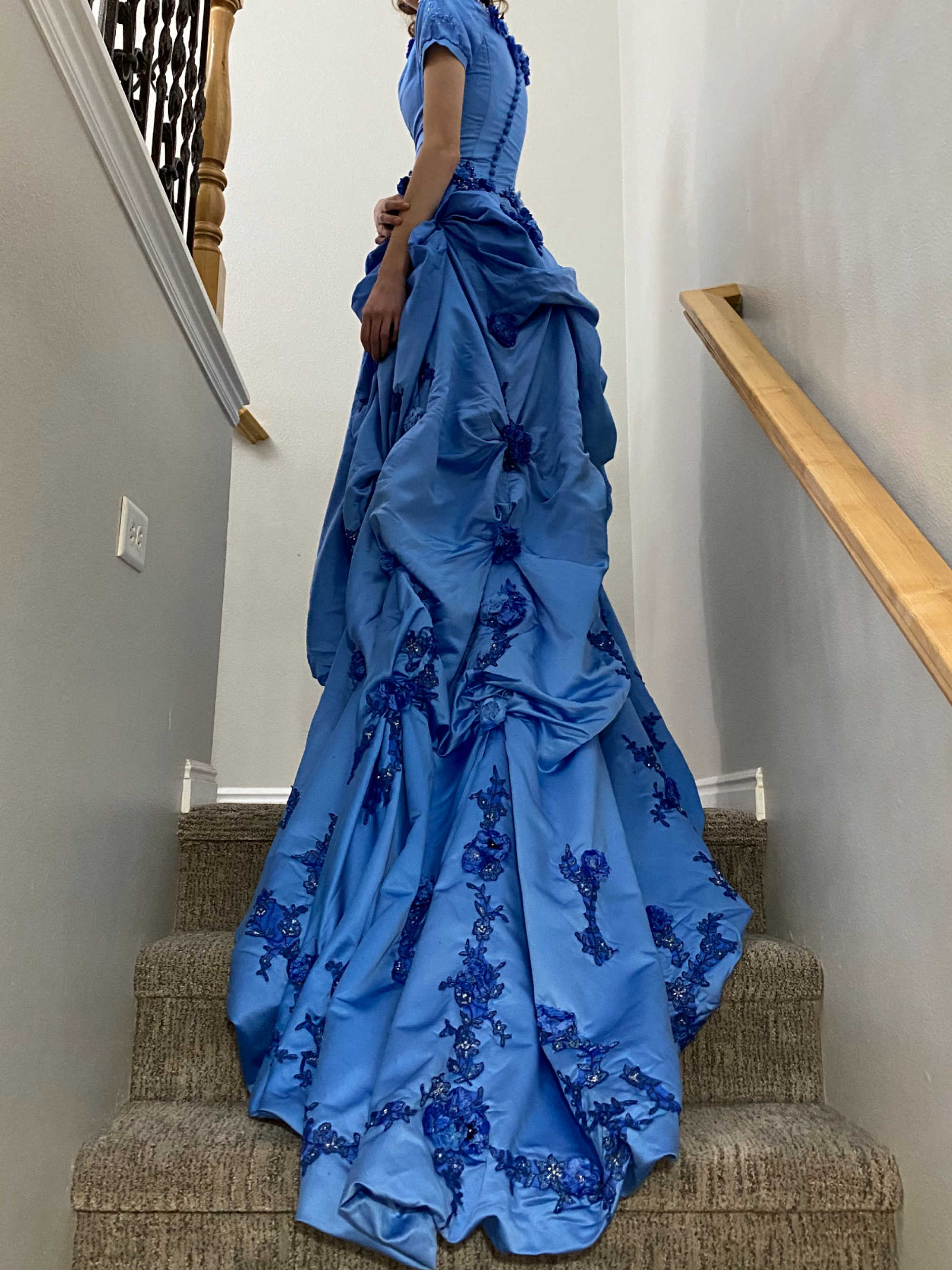 A young woman wearing a secondhand wedding dress from DI that has been dyed blue for Prom. The young woman stands at the top of a staircase to show off the long train of the dress.